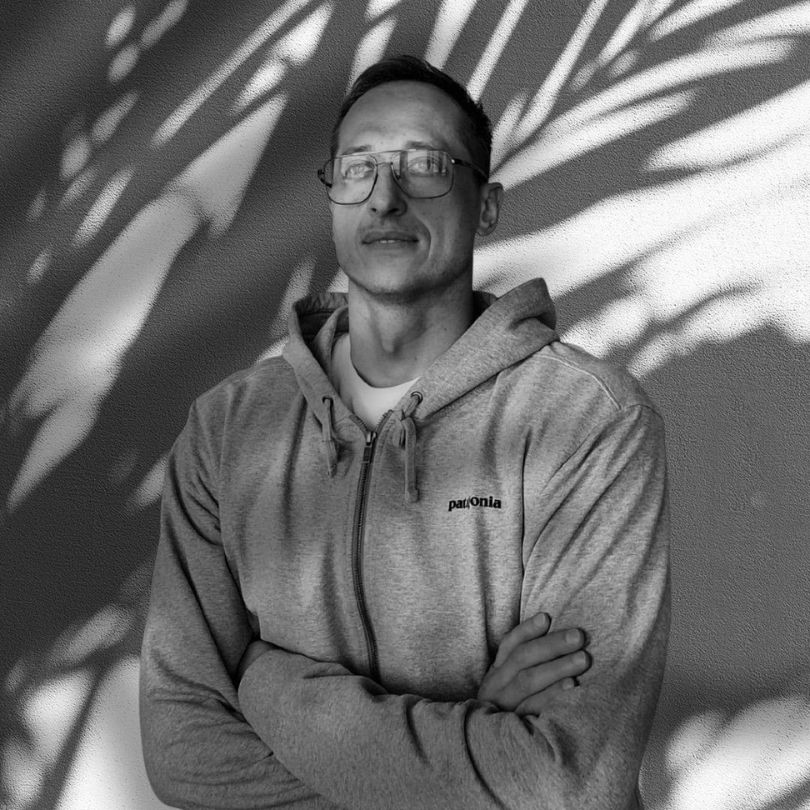 Czarno-biały portret Daniela Kuka, twórcy Optimism Care. Daniel stoi w swobodnym stroju ze skrzyżowanymi rękami, patrząc w przyszłość. Cienie liści na ścianie tworzą artystyczne tło. Zdjęcie ilustruje rolę 'Partnera w Drodze' – przewodnika oferującego wsparcie rówieśnicze oparte na autentycznym doświadczeniu i męskiej stabilności.