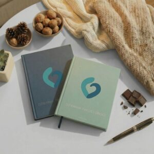 Dwa warianty Dziennika Twojej Drogi (szałwiowy i granatowy) leżące na białym stole w towarzystwie pióra, gorzkiej czekolady, orzechów i ciepłego koca. Przytulna kompozycja zachęcająca do chwili refleksji i pracy z dziennikiem w bezpiecznej atmosferze.
