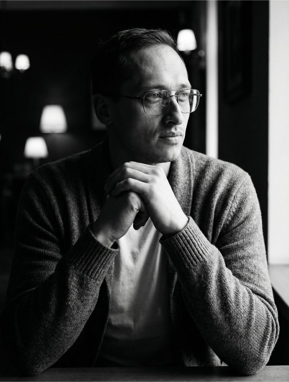 Czarno-biały, refleksyjny portret Daniela Kuka. Założyciel Optimism Care siedzi zamyślony ze splecionymi dłońmi, patrząc w stronę światła. Wizualizacja drogi od mroku uzależnienia do odzyskania wolności.